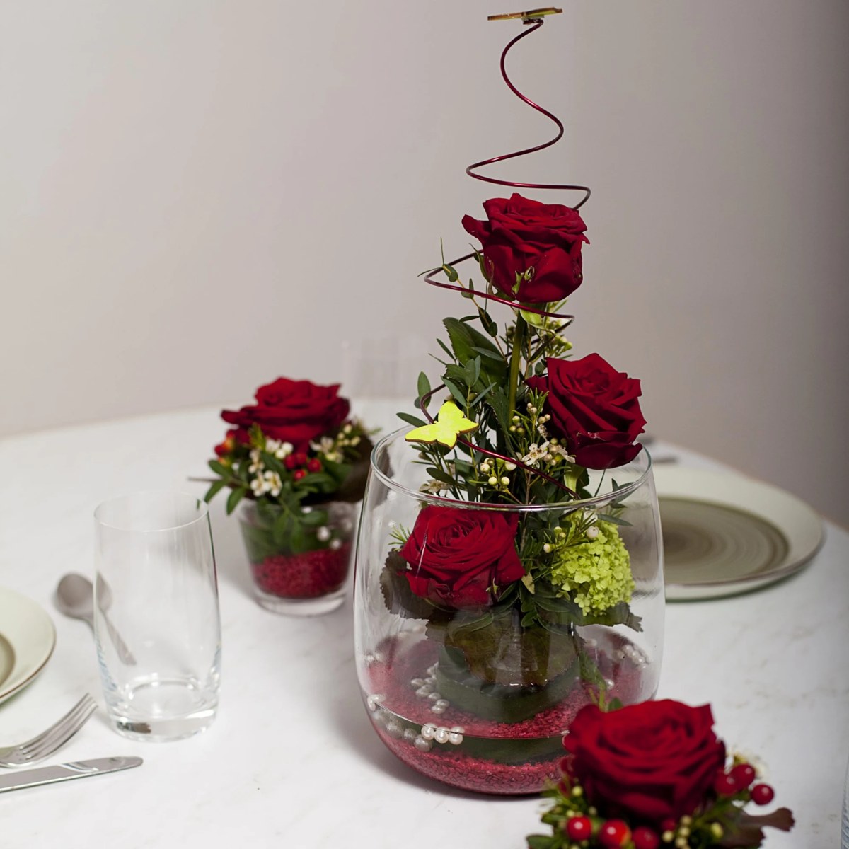 Tischschmuck aus roten Rosen in einer Glasvase mit Drahtspirale und Schmetterling